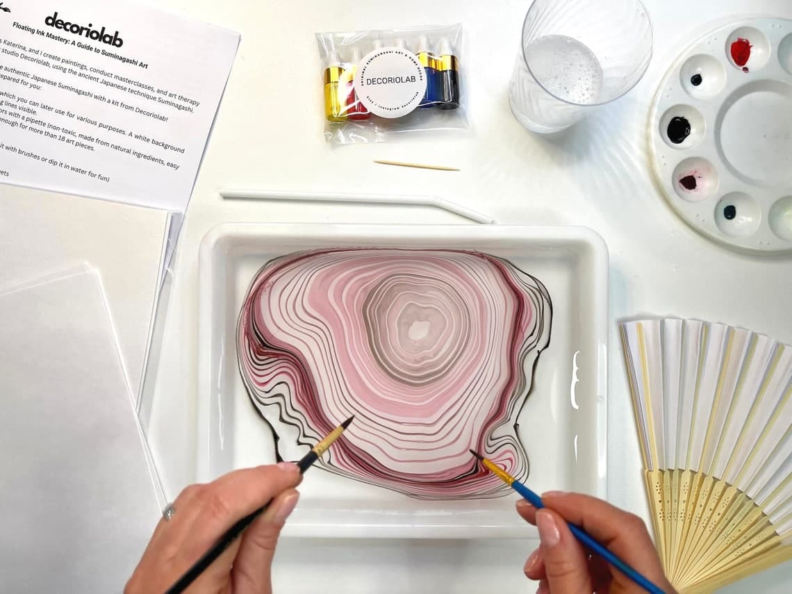 May include: A white tray with a pink and black marbled design in water. Two paintbrushes are being used to create the design. The tray is surrounded by various art supplies, including a palette of paint, a glass of water, and a set of white paper fans.