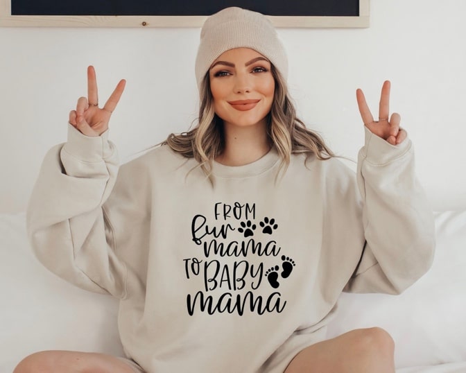 May include: A light beige sweatshirt with the text 'From Fur Mama to Baby Mama' in black. The text is surrounded by paw prints and footprints.