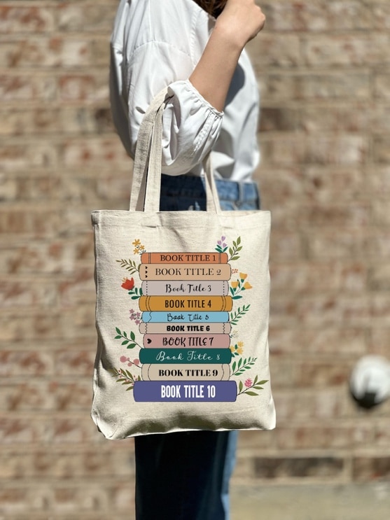 May include: A beige tote bag with a design of stacked books with floral accents. The books have titles that read: 'BOOK TITLE 1', 'BOOK TITLE 2', 'Book Title 3', 'BOOK TITLE 4', 'Back Title 5', 'BOOK TITLE 6', 'BOOK TITLE 7', 'Book Title 8', 'BOOK TITLE 9', and 'BOOK TITLE 10'.