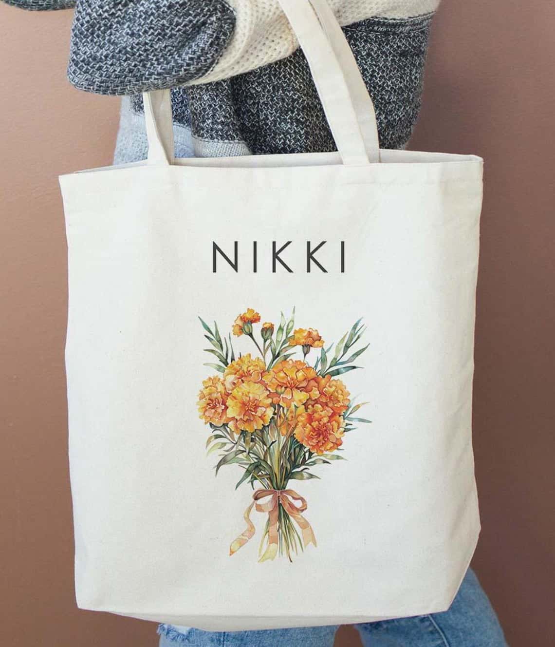 May include: A cream-colored tote bag with a personalized name, 'NIKKI,' printed in a minimalist sans-serif font. A watercolor-style illustration of a bouquet of orange marigolds with green stems and leaves is featured below the name. The bouquet is tied with a light orange ribbon. The bag appears to be made of canvas material.