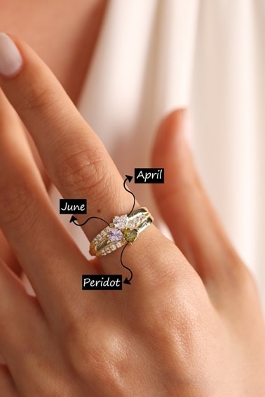 May include: A gold ring with three heart-shaped stones. The stones are a light purple, a light green, and a darker green. The ring is inscribed with the names 'Olivia', 'Emma', and 'June'. The stones are labeled 'April', 'Peridot', and 'June'.