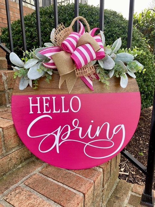 May include: A round wooden door hanger with a pink background and white cursive lettering that says 'Hello Spring'. The door hanger is decorated with a burlap bow with white and pink stripes and a green leafy garland.