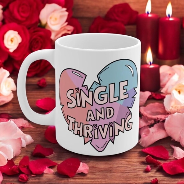 May include: A white coffee mug with a broken heart design. The heart is made up of pink, blue, and purple colors. The text on the mug reads 'Single and Thriving'.