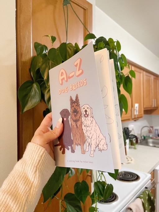 May include: A-Z Dog Breeds coloring book with illustrations of three dogs. The dogs are a brown Labrador Retriever, a German Shepherd, and a Golden Retriever. The book cover is light blue with pink text.