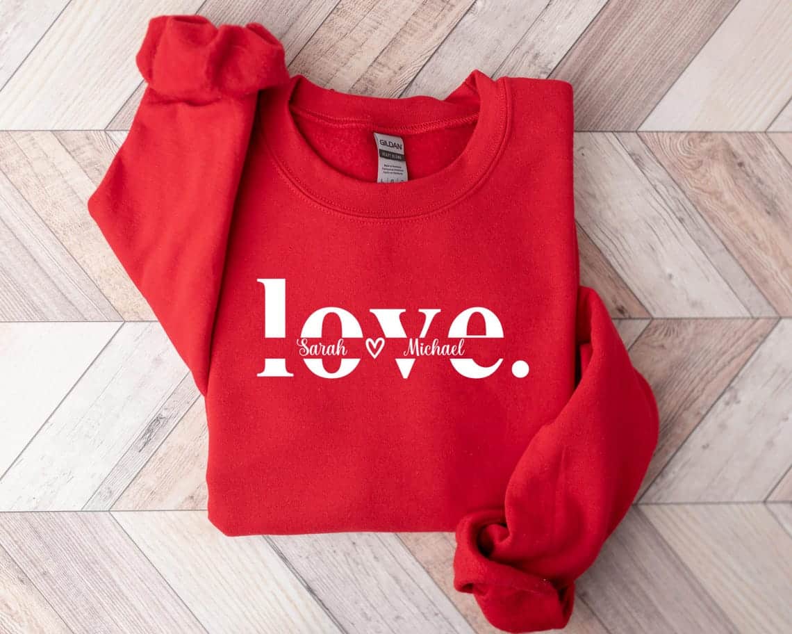 May include: Red sweatshirt with the word 'love' in white letters. The word 'love' is split with the names 'Sarah' and 'Michael' in smaller white letters on either side of the word 'love'.