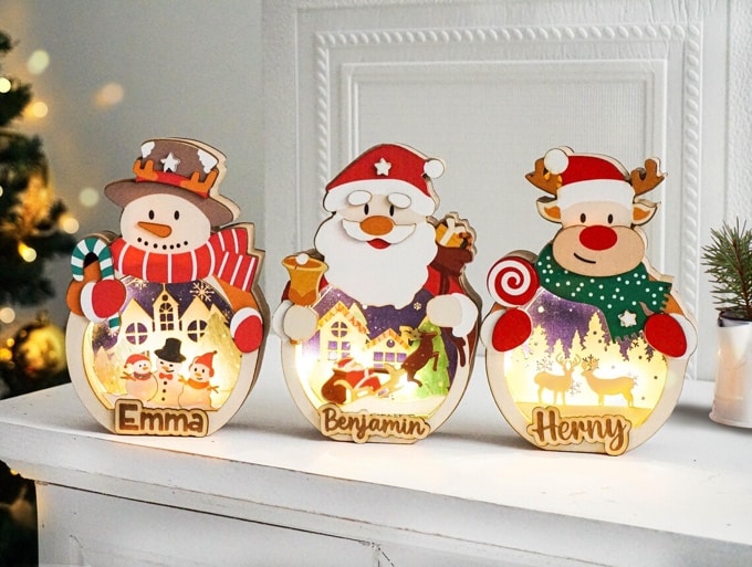 May include: Three illuminated Christmas decorations featuring a snowman, Santa Claus, and a reindeer. Each wooden figure is personalized with a name: Emma, Benjamin, and Herny. The figures are designed with vibrant colors, including red, green, and white, and depict winter scenes with small houses and trees. The decorations are ideal for festive home decor.