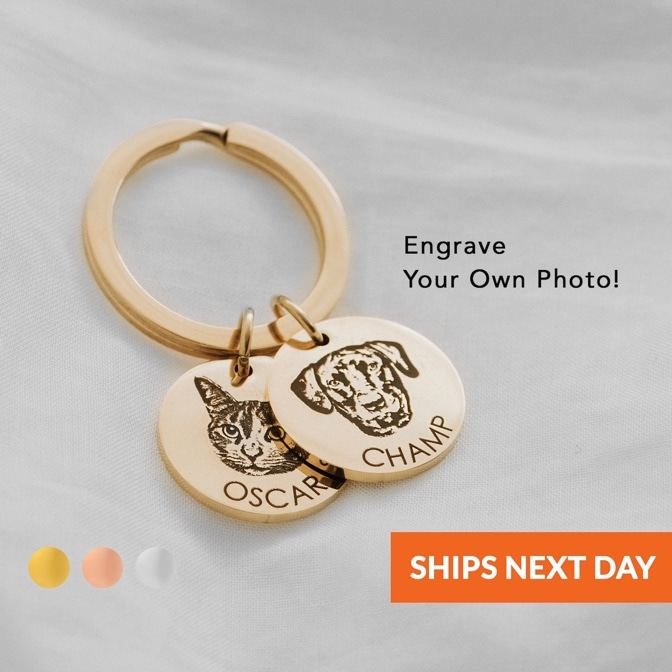 May include: Two gold keychains with engraved photos of a cat and a dog. The cat keychain has the name 'Oscar' engraved on it and the dog keychain has the name 'Champ' engraved on it. The keychains are on a white fabric background. The text 'Engrave Your Own Photo!' is displayed above the keychains. The text 'SHIPS NEXT DAY' is displayed below the keychains. The keychains are available in gold, rose gold, and silver.