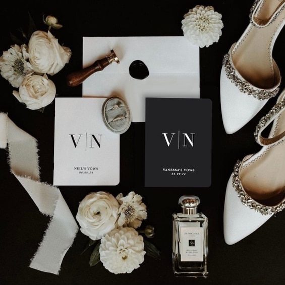 May include: Two white cards with black text on a black background. The text on the cards reads 'V|N' and 'NEIL'S VOWS 06.09.24' on one card and 'V|N' and 'VANESSA'S VOWS 06.09.24' on the other card. There are two white shoes with rhinestones on them, a small bottle of perfume, and white flowers on the background.