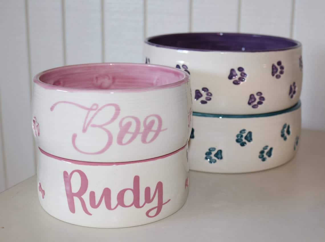 May include: Two white ceramic pet bowls with pink paw prints and pink lettering. The top bowl says 'Boo' and the bottom bowl says 'Rudy'.