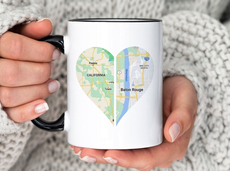 May include: White coffee mug with a black rim and handle. The mug features a heart-shaped design with a map of California on the left side and a map of Baton Rouge, Louisiana on the right side. The text 'California' and 'Baton Rouge' are printed on the maps.