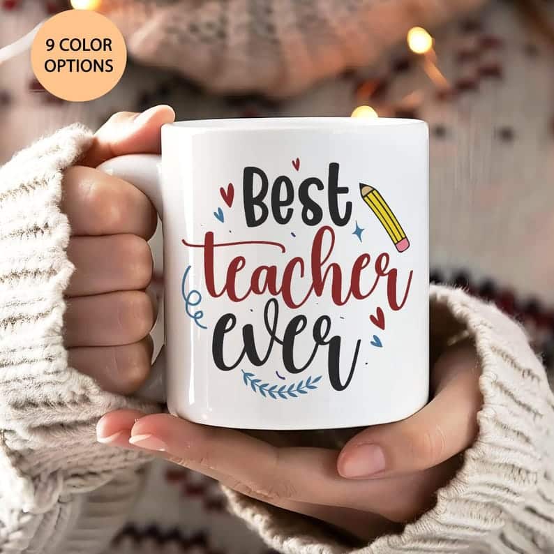May include: White coffee mug with a black and red design that says 'Best teacher ever' with a pencil and hearts. The mug is held in a hand wearing a white sweater.