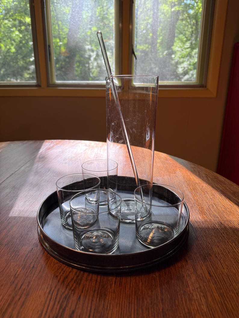 May include: A clear glass cocktail set on a metal tray. The set includes a tall mixing glass, a stirring rod, and several short, round glasses. The tray sits on a wooden table near a window.