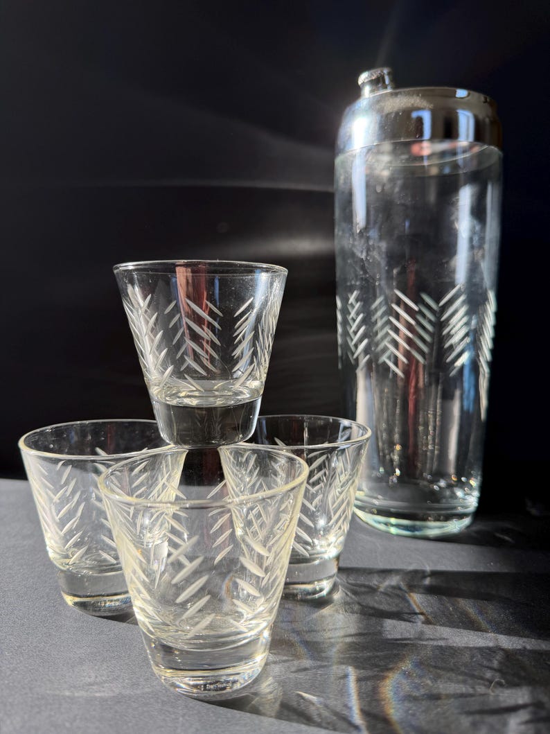 May include: A clear glass cocktail shaker with a silver lid and four shot glasses. The shot glasses and shaker are etched with a repeating leaf pattern. The shot glasses are stacked in a pyramid shape.