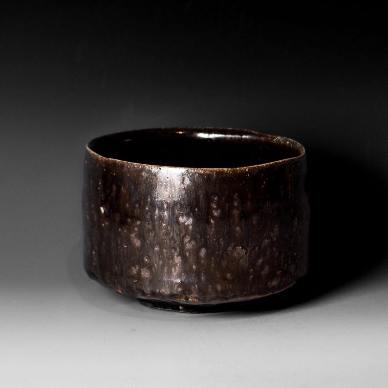 May include: A dark brown ceramic bowl with a wide, cylindrical shape. The bowl has a glossy, textured surface with subtle variations in color. The interior is a deep, dark brown, and the rim is slightly uneven. This handmade bowl is suitable for serving food.