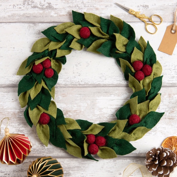 May include: A green and red felt Christmas wreath with red felt berries. The wreath is made of individual felt leaves and is displayed on a white wooden background.