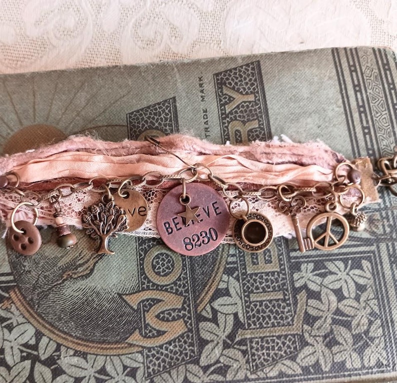 May include: A handmade bracelet with various charms, including a tree, a peace sign, and a disc that says "BELIEVE 8230". The bracelet is adorned with pink ribbons and lace, and is set against a vintage book.