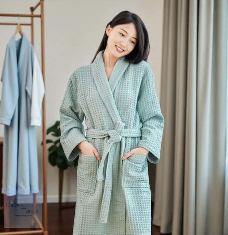 May include: A light green waffle-weave robe with a shawl collar and a matching belt. The robe has two front pockets and is knee-length. Another light blue robe hangs on a wooden rack in the background.