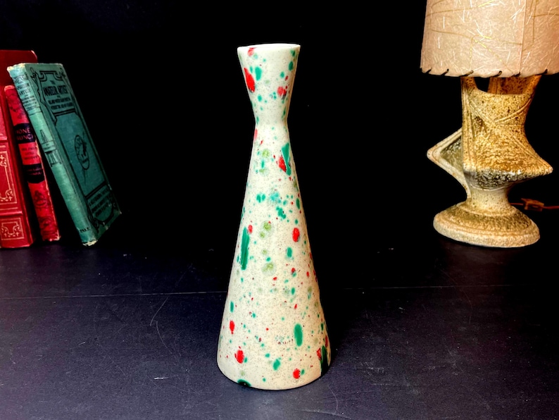 May include: A speckled ceramic vase with a flared base and a narrow neck. The vase is off-white with red and green paint splatters. The vase is approximately 8 inches tall. Decorative home decor.