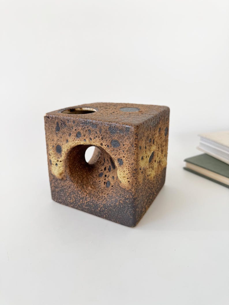 May include: A square, ceramic vase with a textured, speckled brown and tan finish. The vase has a large circular opening on one side and a smaller one on top. The design is geometric and modern, suitable for home decor.