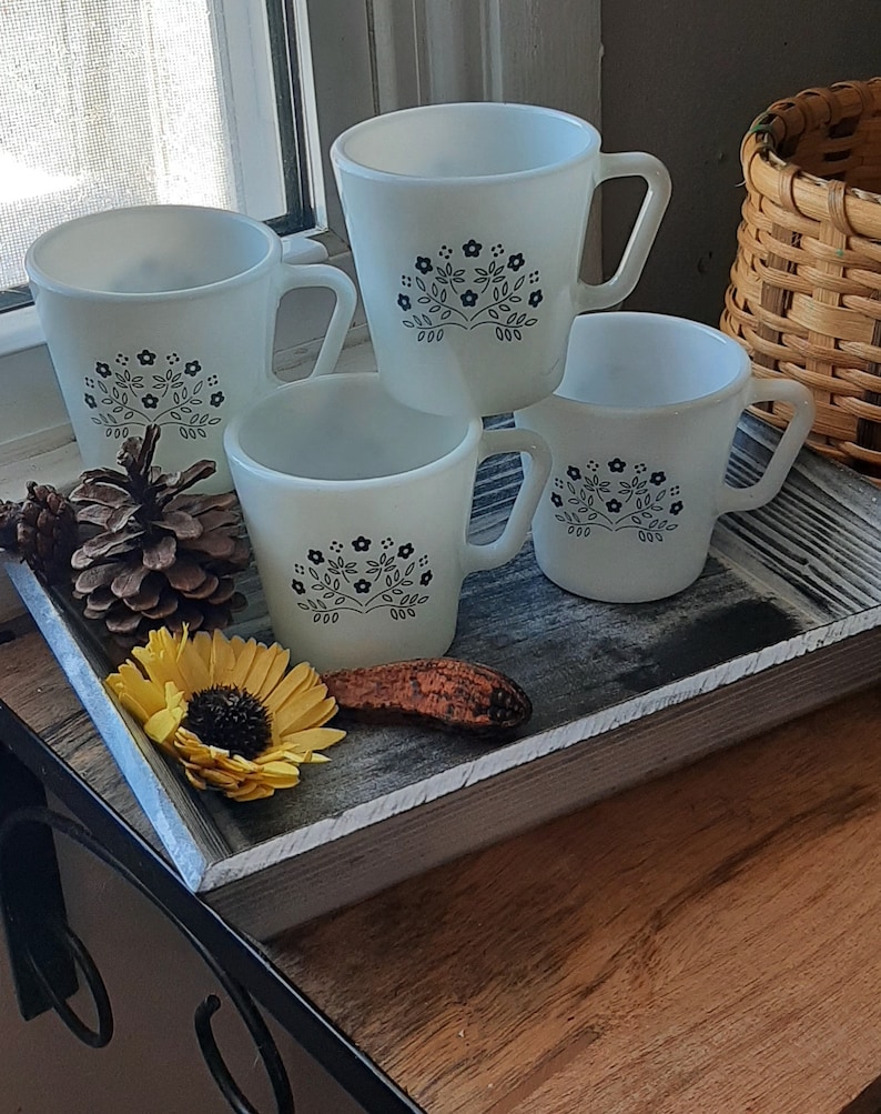 May include: Four white ceramic mugs with black floral designs are stacked on a distressed wooden tray. A yellow sunflower, pine cones, and a decorative piece are also on the tray. The mugs have a classic, vintage aesthetic.