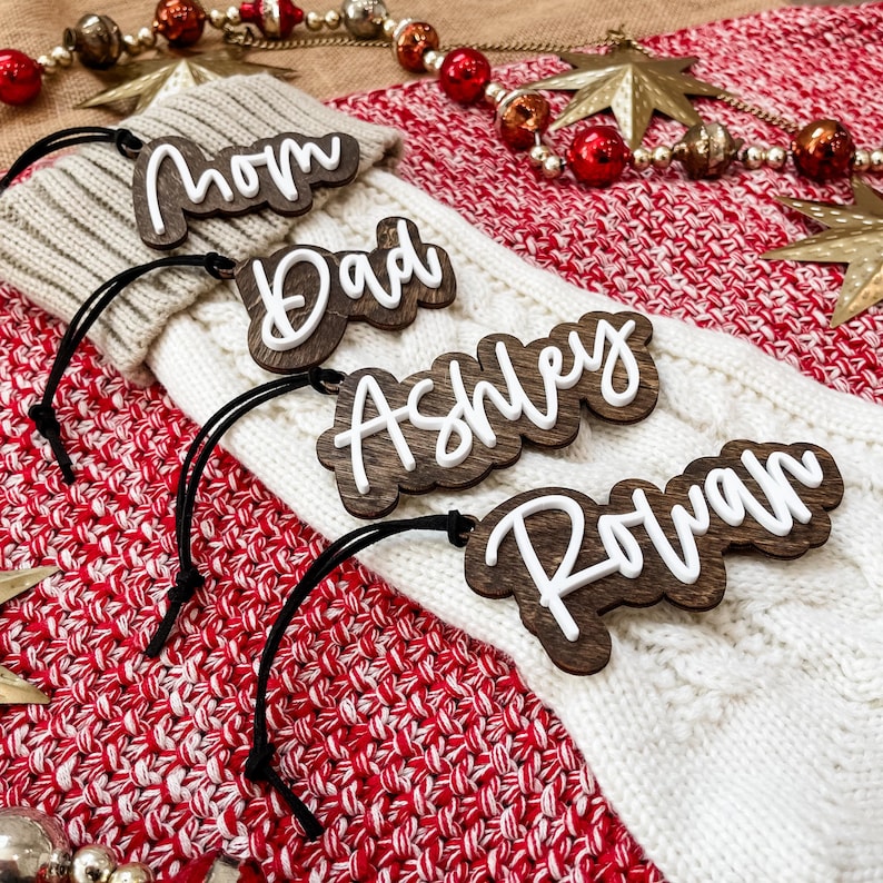 May include: Four wooden ornaments with white lettering spelling out the names "Mom", "Dad", "Ashley", and "Rowan". The ornaments are hanging on a white and red knitted fabric.