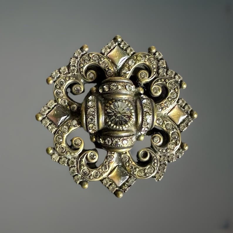 May include: Ornate, vintage-style brooch with a central, round, clear crystal surrounded by smaller clear crystals. The brooch has a detailed, symmetrical design with a metallic, antique gold finish and diamond-shaped accents.