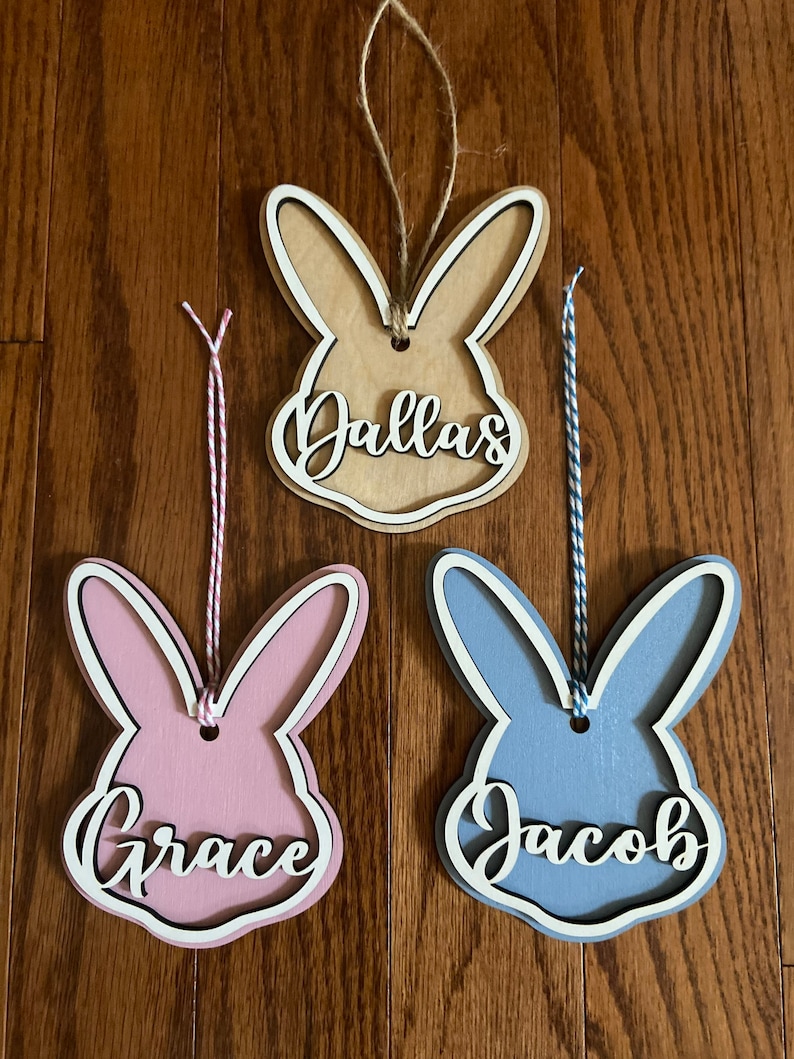 May include: Three wooden bunny-shaped ornaments with white outlines. The ornaments are painted in different colors: brown, pink, and blue. Each ornament has a name written on it: Dallas, Grace, and Jacob.
