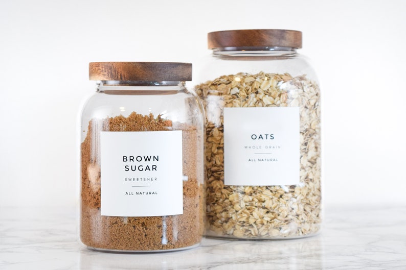 May include: Two glass jars with wooden lids filled with brown sugar and oats. The jars have white labels with black text that reads "Brown Sugar Sweetener All Natural" and "Oats Whole Grain All Natural".