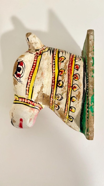 May include: A hand-painted wooden horse head sculpture. The horse is painted white with red, yellow, and black decorative accents. The sculpture is mounted on a green and brown wooden base, showcasing a rustic aesthetic.