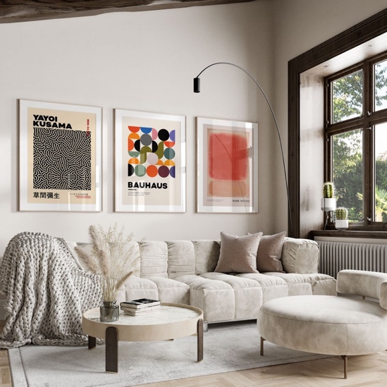 May include: A living room interior with a white sofa, a round coffee table, and a white ottoman. Three framed posters hang on the wall behind the sofa. The posters feature abstract designs and text, including "YAYOI KUSAMA" and "BAUHAUS".