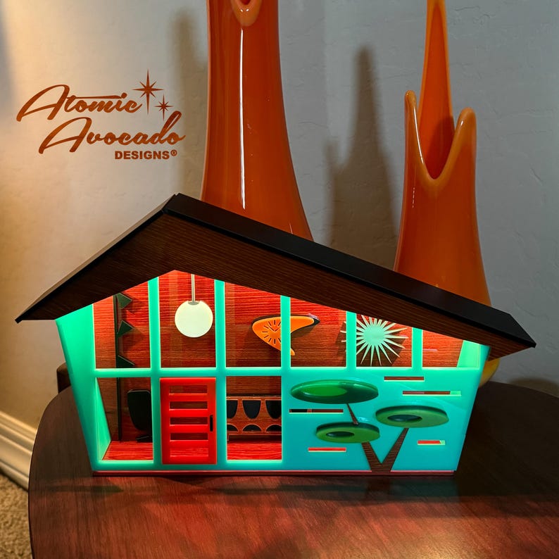 May include: A teal blue and red mid-century modern house shaped light with a wood roof and a white light bulb hanging from the ceiling. The light is lit and the interior is illuminated with a warm glow. The text "Atomic Avocados Designs" is visible in the top left corner of the image.