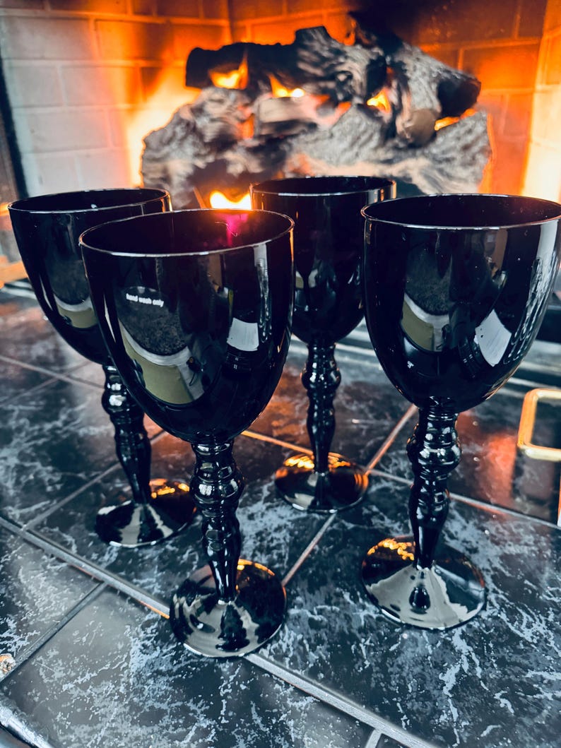 May include: Four black wine glasses with ornate stems are arranged on a tiled surface. The glasses have a dark, reflective quality, and the background features a fireplace with a warm, glowing fire.