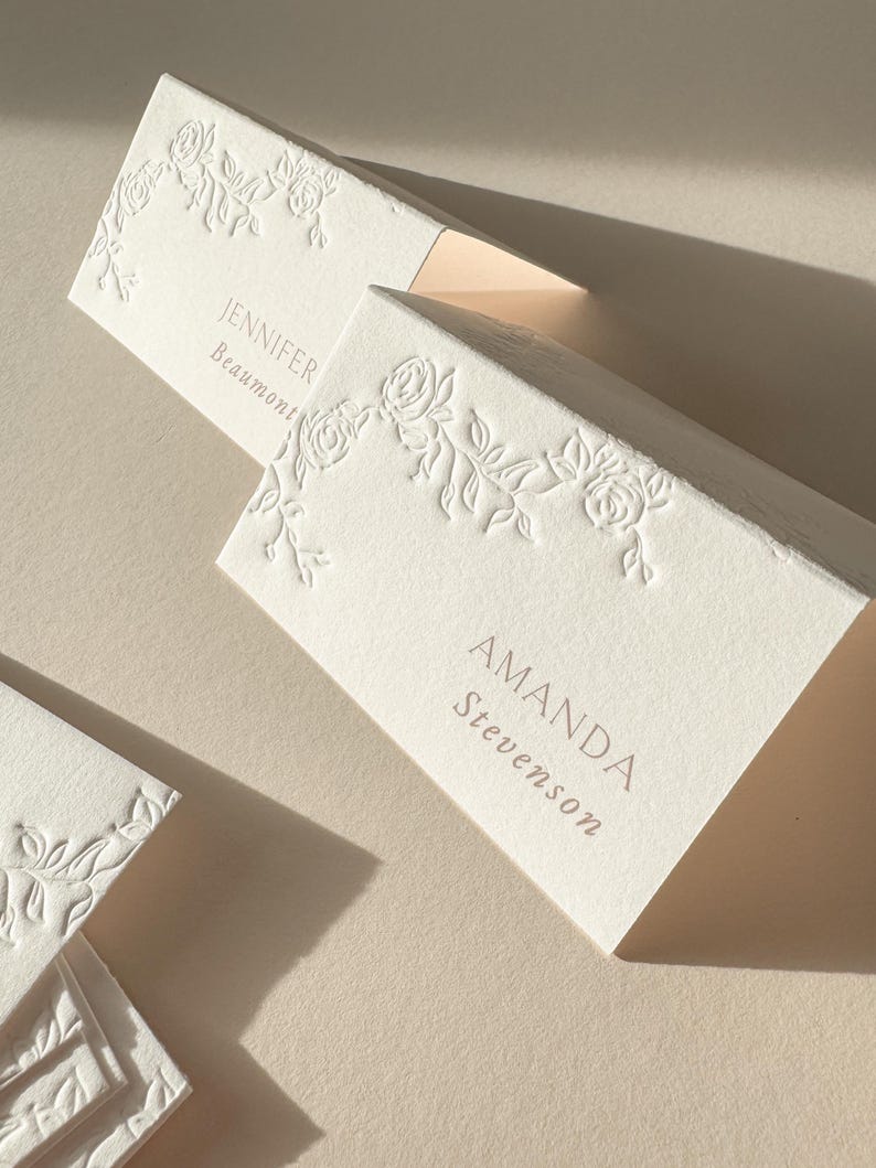 May include: Ivory place cards with embossed floral designs. The cards feature names and surnames in a soft, muted font. The cards are arranged on a neutral surface, with soft lighting creating shadows.