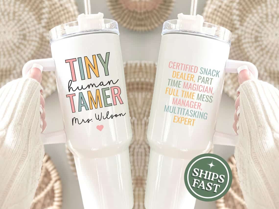May include: Two white insulated tumblers with lids and handles. One tumbler reads "TINY human TAMER Mrs. Wilson" in colorful text with a heart. The other reads "CERTIFIED SNACK DEALER, PART TIME MAGICIAN, FULL TIME MESS MANAGER, MULTITASKING EXPERT."