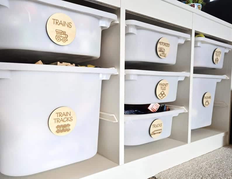 May include: White plastic storage bins with wooden labels on the front. The labels have different toy categories written on them, including "Trains", "Train Tracks", "Motorized", "Magnet Tiles", "Trucks", and "Cars".