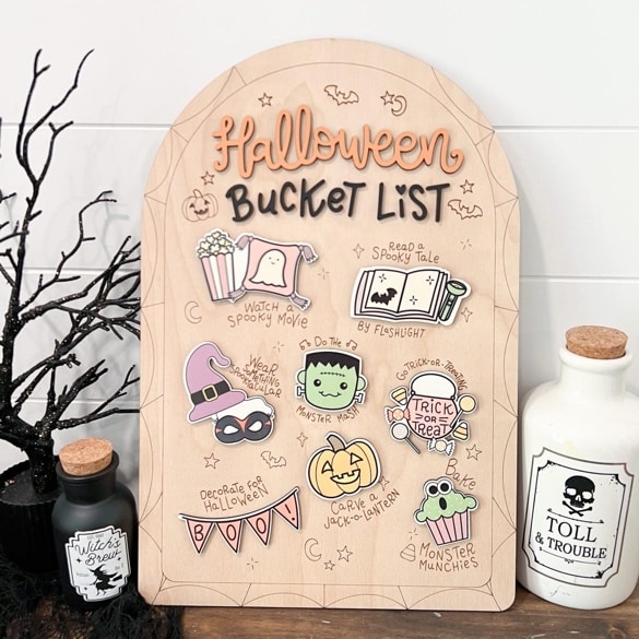 May include: Wooden Halloween bucket list sign with decorative illustrations and the words "Halloween Bucket List". Includes images of a ghost, popcorn, a book, a monster, a pumpkin, and a cupcake. Includes the text "Witch's Brew" and "Toll & Trouble".
