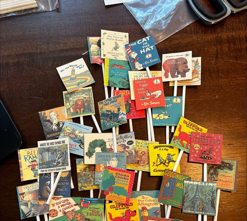 May include: A collection of colorful book-themed cake toppers featuring popular children's books such as "Where the Wild Things Are", "The Cat in the Hat", "Goodnight Moon", "Madeline", "Click, Clack, Moo: Cows That Type", and "Corduroy".