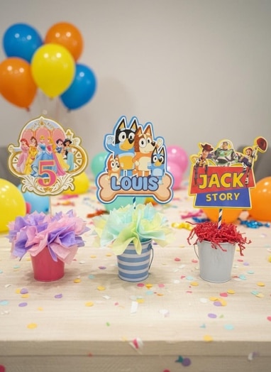 May include: Colorful birthday party decorations featuring character cutouts, balloons, and decorative buckets. The cutouts include Disney princesses, Bluey characters, and Toy Story characters. The buckets hold tissue paper flowers and shredded paper.